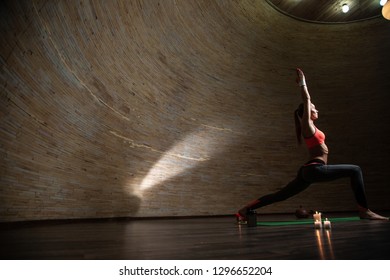 Young Beautiful Woman Standing On The Yoga Mat In Dark Empty Room And Practicing The Warrior Pose With Candles On The Floor. Copy Space On The Left Side