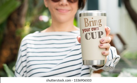 A young beautiful woman standing in the outdoor garden, she hold a reusable insulated stainless tumbler glass with the word "Bring your own cup" to show awareness of 'Say no to plastic', Copy space.