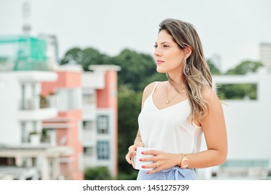 Young beautiful woman standing outdoors and looking away with pensive look while drinking coffee