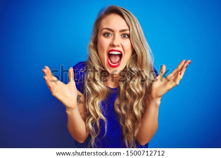Young beautiful woman standing over blue isolated background celebrating crazy and amazed for success with arms raised and open eyes screaming excited. Winner concept