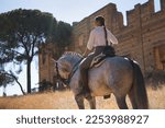 Young and beautiful woman standing, riding her horse on a country road next to an abandoned and ruined building, under the rays of the sun. Concept horse riding, animals, dressage, horsewoman.