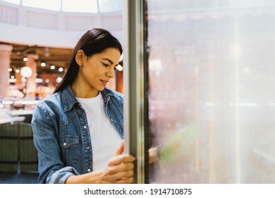 Young Beautiful Woman Stands Opening Glass Freezer Door In Grocery Store And Selects Products.