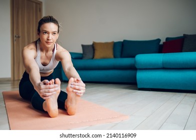 Young Beautiful Woman Stretching To Touch Toes While Sitting On Yoga Mat And Training At Home On The Floor. Paschimottanasana Or Seated Forward Bend Pose.