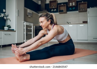 Young Beautiful Woman Stretching To Touch Toes While Sitting On Yoga Mat And Training At Home On The Floor. Paschimottanasana Or Seated Forward Bend Pose.