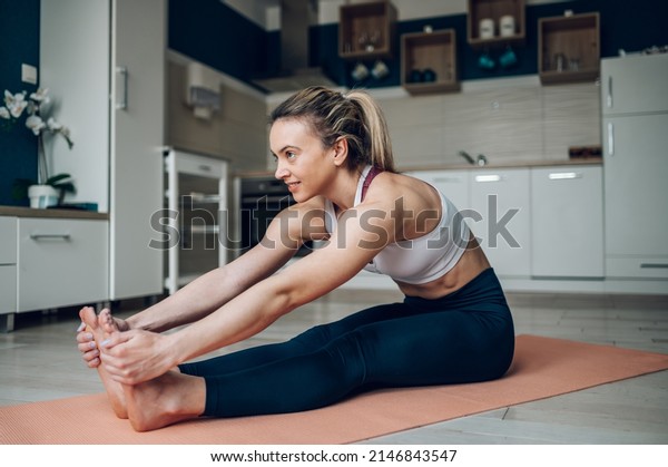 Young beautiful woman stretching to\
touch toes while sitting on yoga mat and training at home on the\
floor. Paschimottanasana or seated forward bend\
pose.