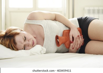 Young Beautiful Woman Suffering Stomach Cramps On Belly Holding With Hands Hot Water Bottle Against Tummy In Period Pain Lying On Bed At Home In Menstruation Hurt Concept