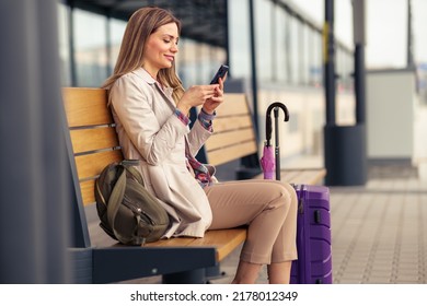 A young and beautiful woman with a suitcase and a backpack is sitting at the train station waiting for a train while using a mobile phone.