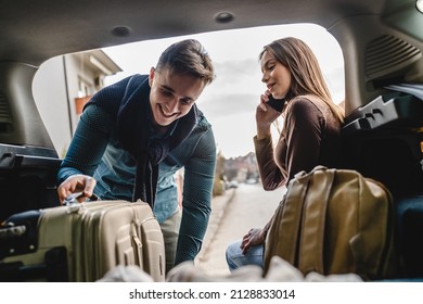 Young beautiful woman talking on the phone making a call from the back of her car sitting in the trunk while her boyfriend or husband it taking luggage baggage and other belongings from the car travel - Shutterstock ID 2128833014
