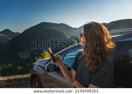 Young beautiful woman traveling by car in the mountains using smartphone at sunset, summer vacation and adventure