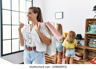 Young Beautiful Woman With Two Daughters Holding Shopping Bags At Retail Shop Pointing Thumb Up To The Side Smiling Happy With Open Mouth 