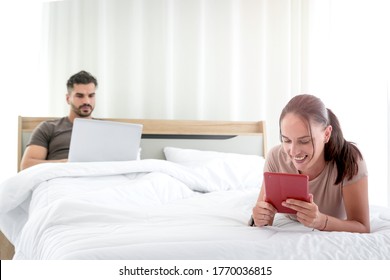 Young Beautiful Woman Using Digital Tablet Lying On Bed In Bedroom With Man Using Laptop Computer As Background, Searching Information And Shopping, Social Media For Online Business, Work Form Home