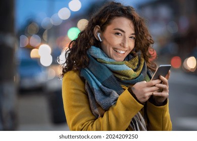 Young beautiful woman using mobile phone at dusk in the city while listening music through earphones. Happy woman using smartphone to do a video call on a winter day while looking at camera.