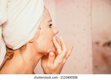 Young beautiful woman using a moisturizing facial mask after taking a bath. Pretty attractive girl in a towel on her head stands in front of a mirror in a home bathroom. Daily hygiene and skin care - Shutterstock ID 2125800308