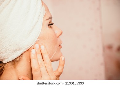 Young beautiful woman using a moisturizing facial mask after taking a bath. Pretty attractive girl in a towel on her head stands in front of a mirror in a home bathroom. Daily hygiene and skin care