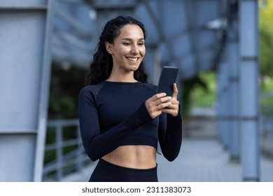 Young beautiful woman using phone, sportswoman smiling and resting after running and doing active physical exercises, Hispanic woman with curly hair in tracksuit. - Powered by Shutterstock