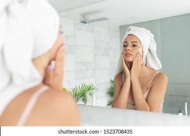 Young Beautiful Woman Using Skin Face Cream Moisturizing Lotion After Taking Bath. Pretty Attractive Girl Wearing Towel On Head Standing Front Of Mirror In Home Bathroom. Daily Hygiene And Skincare