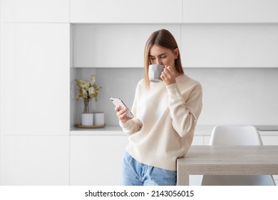 A young beautiful woman using a smartphone and drinking morning tea or coffee in the kitchen at home - Powered by Shutterstock