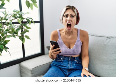 Young Beautiful Woman Using Smartphone Typing Message Sitting On The Sofa Angry And Mad Screaming Frustrated And Furious, Shouting With Anger. Rage And Aggressive Concept. 