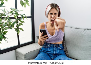 Young Beautiful Woman Using Smartphone Typing Message Sitting On The Sofa Pointing Displeased And Frustrated To The Camera, Angry And Furious With You 