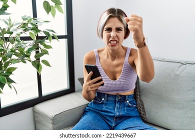 Young Beautiful Woman Using Smartphone Typing Message Sitting On The Sofa Angry And Mad Raising Fist Frustrated And Furious While Shouting With Anger. Rage And Aggressive Concept. 