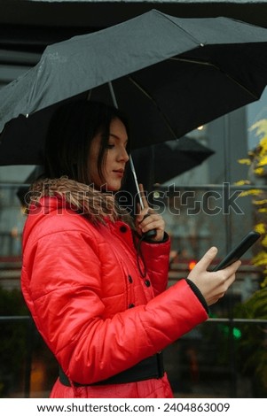 Young beautiful woman using smartphone while holding a umbrella and walking in a city on a rainy day
