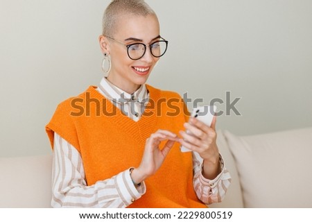 Young  beautiful woman with very short hair sitting  on sofa and using smartphone, texting message or surfing internet while relaxing at home. Female with cellphone