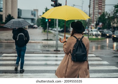 Young Beautiful Woman Walking On The Crossroad During Rain And Holding A Yellow Umbrella Dressed In A Coat