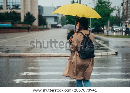 Young beautiful woman walking on the crossroad during rain and holding a yellow umbrella dressed in a coat