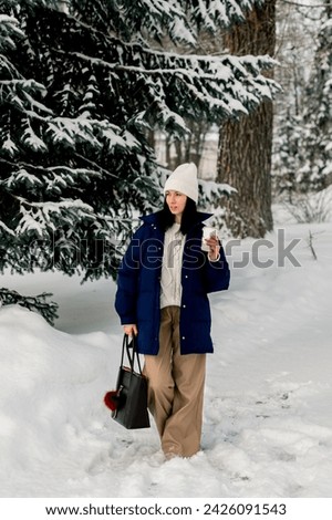 A young beautiful woman is walking in a winter park with a glass of hot coffee or tea in her hands.