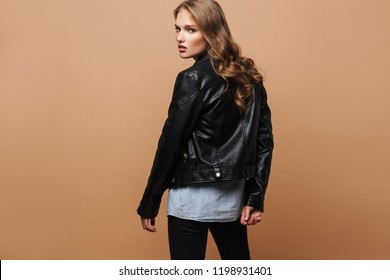 Young Beautiful Woman With Wavy Hair In Black Leather Jacket Thoughtfully Looking In Camera From Back Over Beige Background