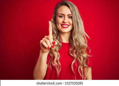 Young beautiful woman wearing basic t-shirt standing over red isolated background showing and pointing up with finger number one while smiling confident and happy.