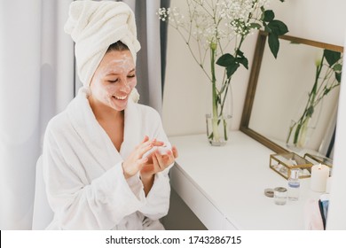 Young beautiful woman wearing bathrobe and towel on her hair applying moistrizing cream on her face. Skin care morning rituals. Beauty routine.  - Powered by Shutterstock