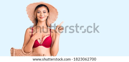 Young beautiful woman wearing bikini and hat holding summer wicker handbag surprised with an idea or question pointing finger with happy face, number one 
