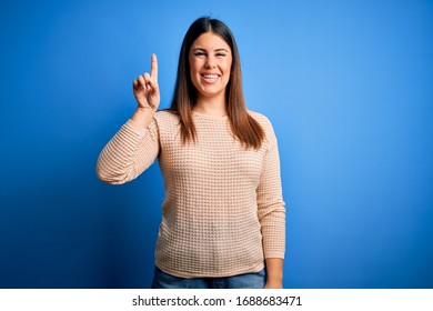 Young beautiful woman wearing casual sweater over blue background showing and pointing up with finger number one while smiling confident and happy. - Shutterstock ID 1688683471