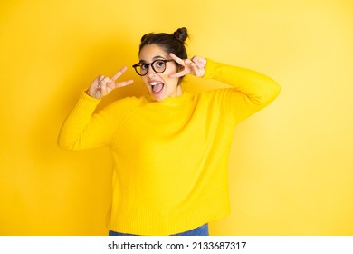 Young Beautiful Woman Wearing Casual Sweater Over Isolated Yellow Background Doing Peace Symbol With Fingers Over Face, Smiling Cheerful Showing Victory