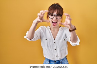 Young beautiful woman wearing casual shirt over yellow background shouting frustrated with rage, hands trying to strangle, yelling mad 