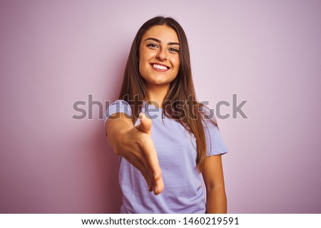 Young beautiful woman wearing casual t-shirt standing over isolated pink background smiling friendly offering handshake as greeting and welcoming. Successful business.
