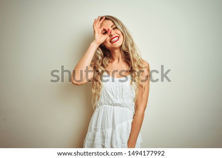 Young beautiful woman wearing casual dress standing over white isolated background doing ok gesture with hand smiling, eye looking through fingers with happy face.