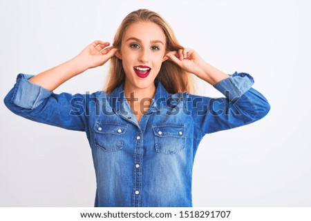 Young beautiful woman wearing casual denim shirt standing over isolated white background Smiling pulling ears with fingers, funny gesture. Audition problem