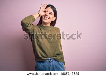 Young beautiful woman wearing casual sweater standing over isolated pink background doing ok gesture with hand smiling, eye looking through fingers with happy face.