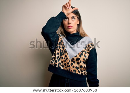 Young beautiful woman wearing casual sweatshirt standing over isolated white background making fun of people with fingers on forehead doing loser gesture mocking and insulting.
