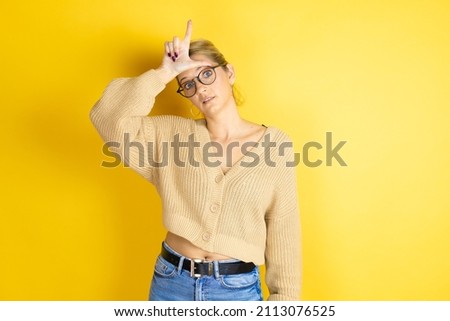 Young beautiful woman wearing casual sweater over isolated yellow background making fun of people with fingers on forehead doing loser gesture mocking and insulting.