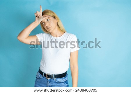 Young beautiful woman wearing casual t-shirt over isolated blue background making fun of people with fingers on forehead doing loser gesture mocking and insulting.