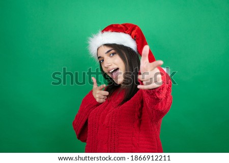Young beautiful woman wearing Christmas Santa hat over isolated green background pointing to you and the camera with fingers, smiling positive and cheerful