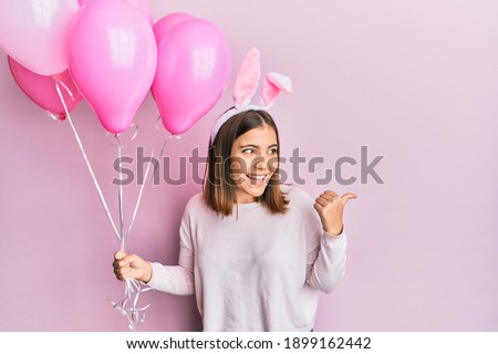 Young beautiful woman wearing cute easter bunny ears and holding balloons pointing thumb up to the side smiling happy with open mouth 