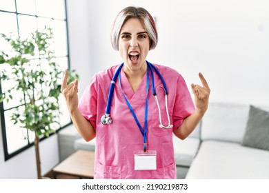 Young Beautiful Woman Wearing Doctor Uniform And Stethoscope Shouting With Crazy Expression Doing Rock Symbol With Hands Up. Music Star. Heavy Concept. 