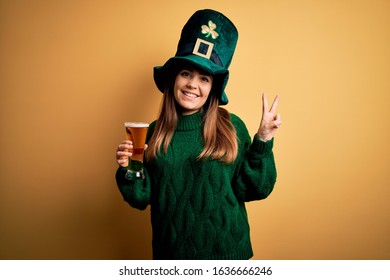 Young beautiful woman wearing green hat drinking glass of beer on saint patricks day smiling looking to the camera showing fingers doing victory sign. Number two.