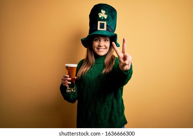 Young beautiful woman wearing green hat drinking glass of beer on saint patricks day smiling with happy face winking at the camera doing victory sign with fingers. Number two.