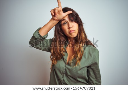 Young beautiful woman wearing green shirt standing over grey isolated background making fun of people with fingers on forehead doing loser gesture mocking and insulting.