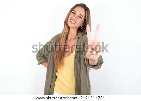 young beautiful woman wearing green overshirt over white background smiling and looking happy, carefree and positive, gesturing victory or peace with one hand